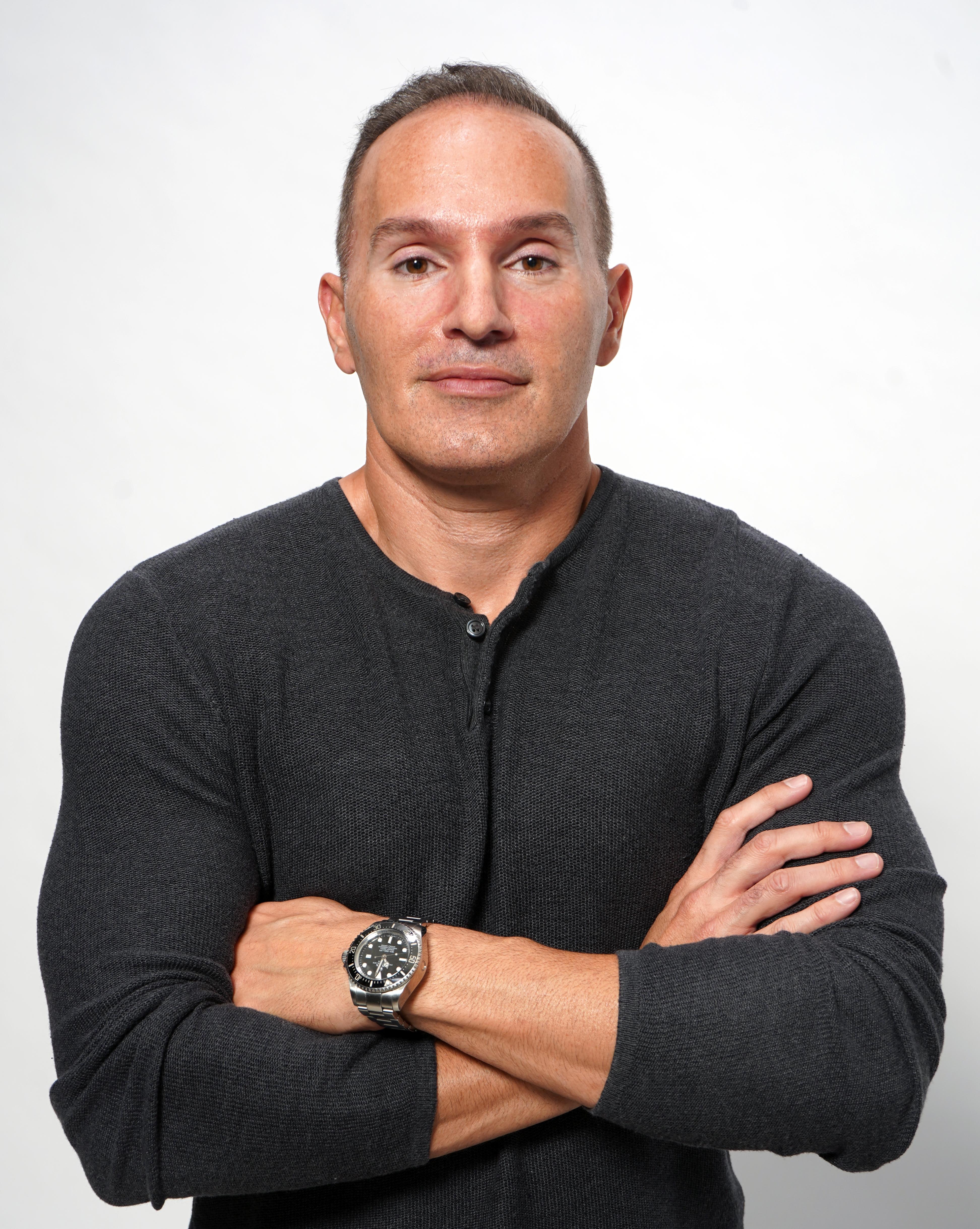 Portrait of a Verano executive wearing a dark long-sleeve shirt with arms crossed against a light gray background.